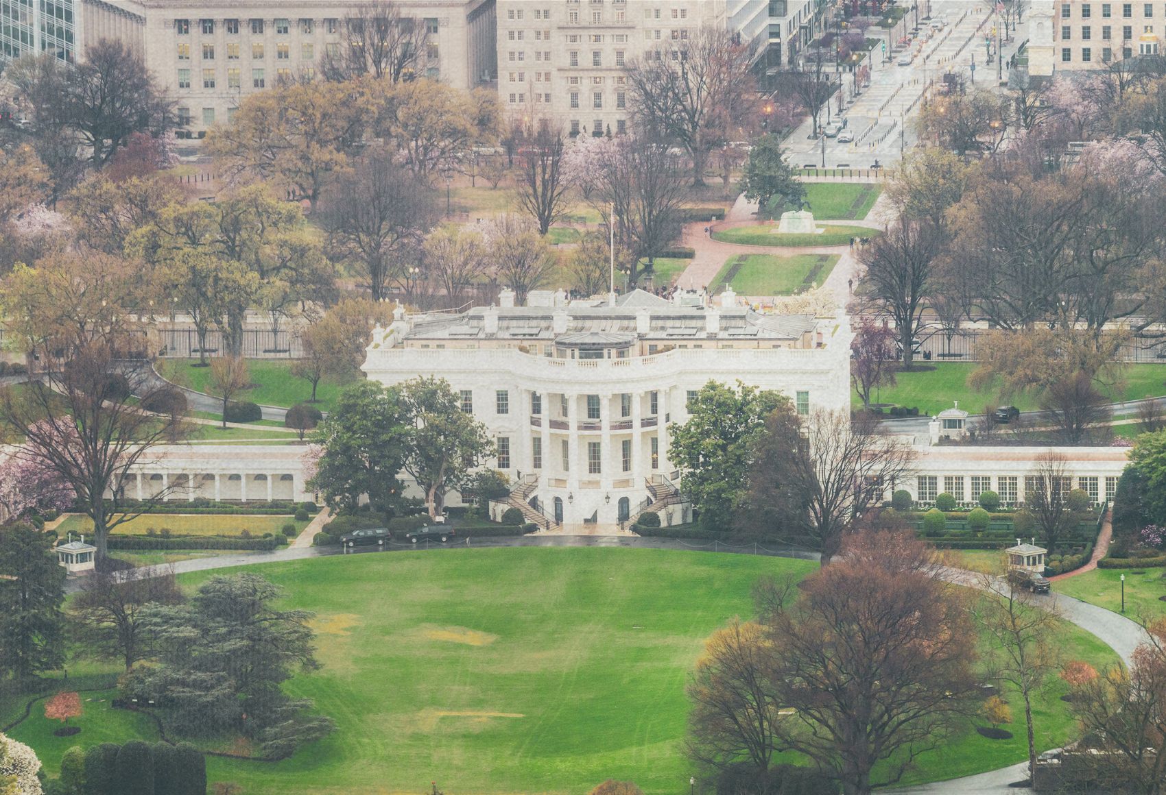 Aerial view of the White House surrounded by trees and greenery, with nearby buildings and streets visible in the background.
