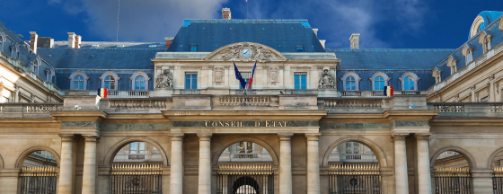 Facade of France's Conseil d'État, featuring stone archways, ornate sculptures, a clock, and French and European Union flags beneath a cloudy blue sky.